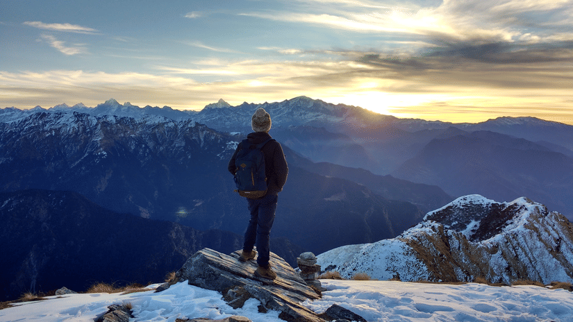 A person standing atop a snowy mountain peak, overlooking a breathtaking sunset sky and distant ranges