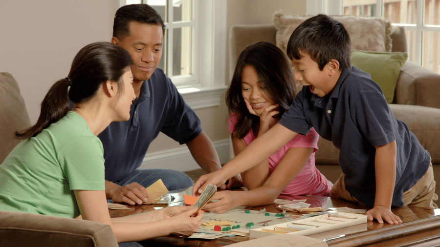 Family gathered around a table indoors, engaging in a board game with colorful tiles and focused expressions, suggesting a moment of shared activity