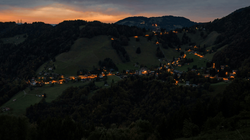 A panoramic view of a valley at dusk or dawn, with rolling hills, scattered lights from buildings, and a colourful sky