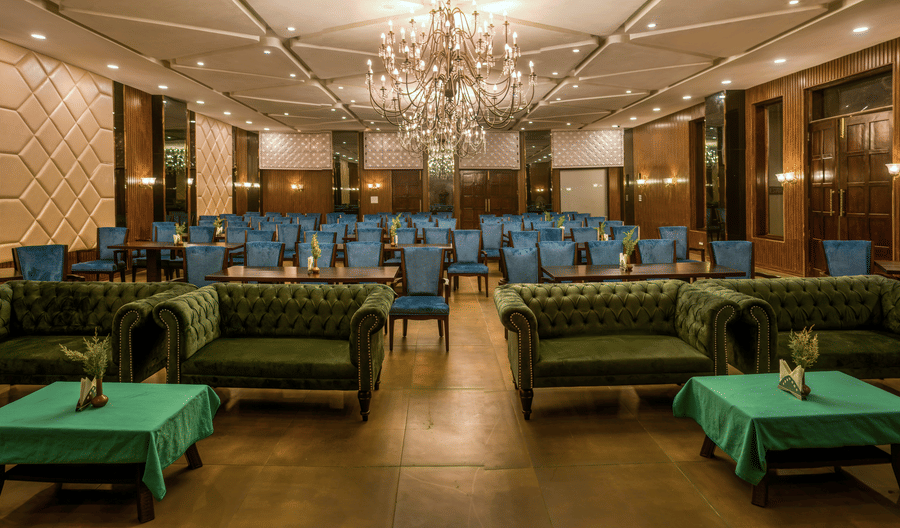 A large dining hall at Digantaa Resorts in Mukteshwar featuring high ceilings, chandeliers, and numerous tables with green tablecloths and matching chairs, ready for guests