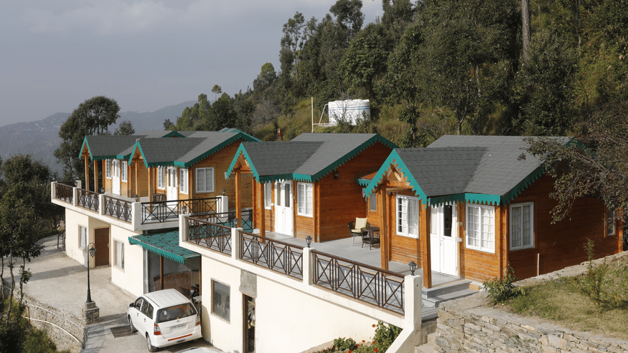 A scenic view of multiple charming cottages and bungalows at Digantaa Resort in Mukteshwar featuring grey roofs, set on a verdant hillside with trees in the background