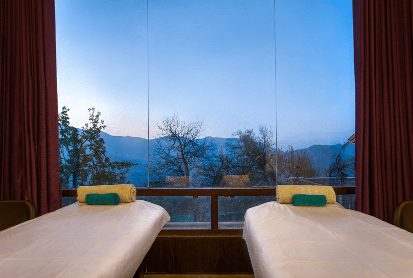 Two massage beds in a spa room with large windows  at Digantaa Resort in Mukteshwar providing a tranquil view of the mountains
