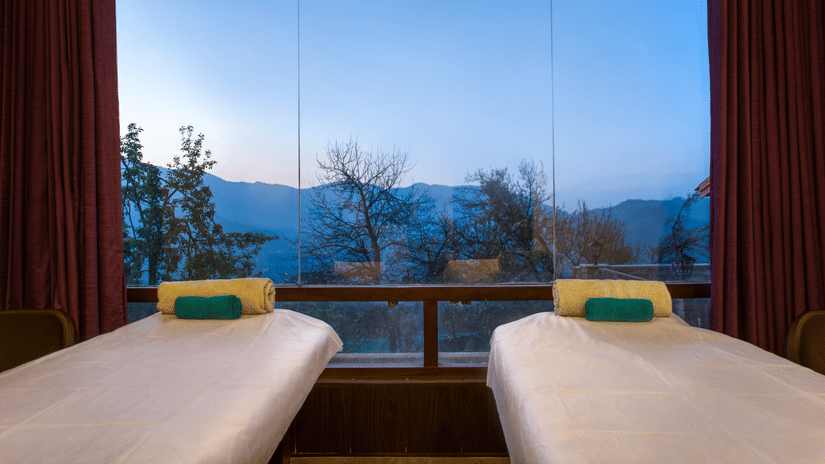 Two massage beds in a spa room with large windows  at Digantaa Resort in Mukteshwar providing a tranquil view of the mountains