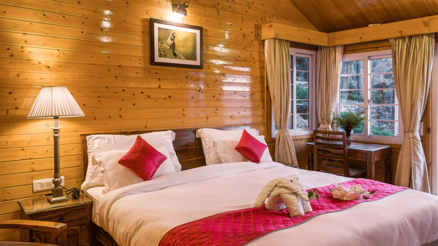  A serene bedroom with light wooden walls and ceiling at Digantaa Resorts in Mukteshwar A large bed with a white comforter and a red decorative runner is prominently featured. A lamp on a bedside table illuminates the left side of the bed